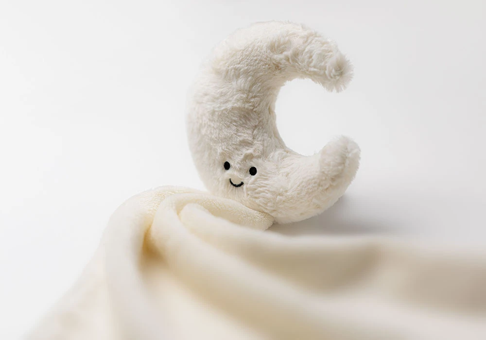 Amuseables Moon sitting at the corner of a soft creme blanket
