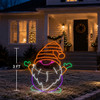 A 3-foot-tall LED neon gnome yard decoration displayed on the front lawn of a suburban home at dusk. The gnome features an orange striped hat, white beard, purple body, green mittens and boots, and a pink nose, glowing brightly against the winter landscape. Warm porch lights, a holiday wreath, and a lit Christmas tree visible through the window create a cozy, festive outdoor setting.