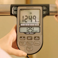 Hands holding the Omron BF511 handlebar sensors during a body composition analysis, with results visible on the screen.