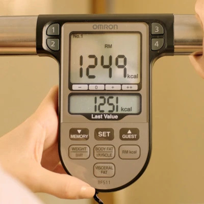 Hands holding the Omron BF511 handlebar sensors during a body composition analysis, with results visible on the screen.