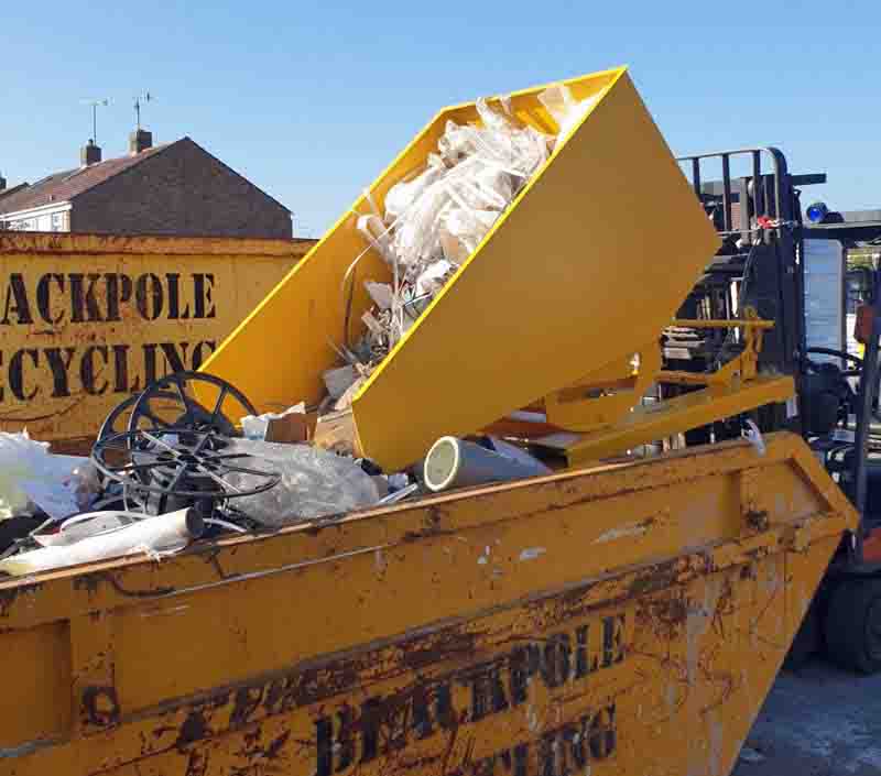 Un contenedor basculante amarillo acoplado a una carretilla elevadora que se inclina hacia adelante mientras vacía su contenido en un contenedor.