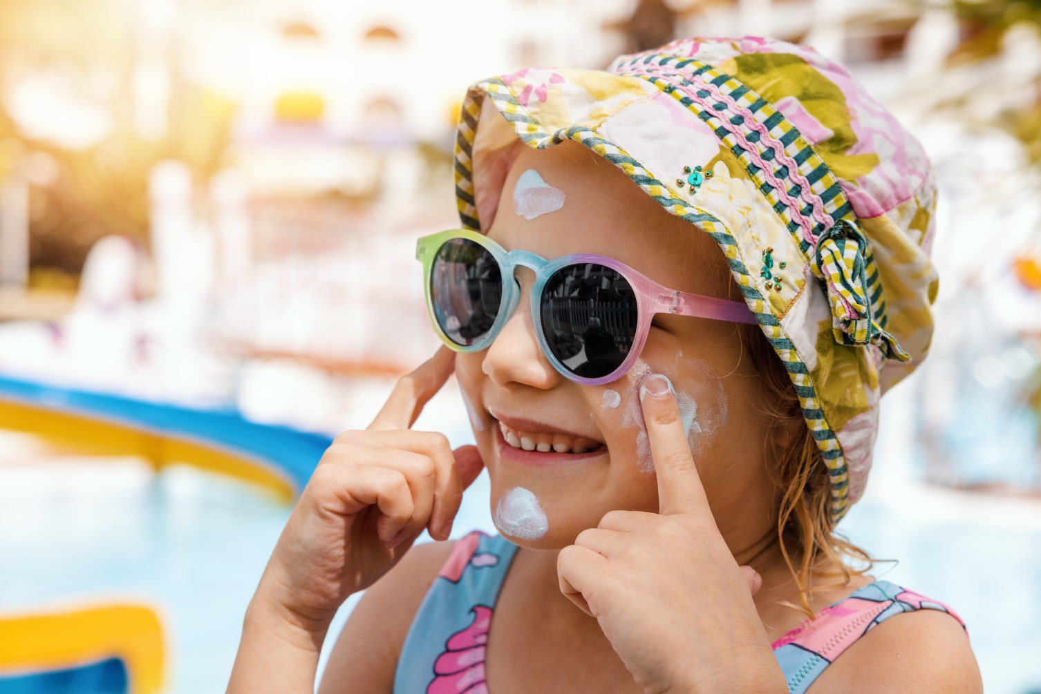 A young girl dressed in summer clothes, including a summer hat and sunglasses, applying suncream.