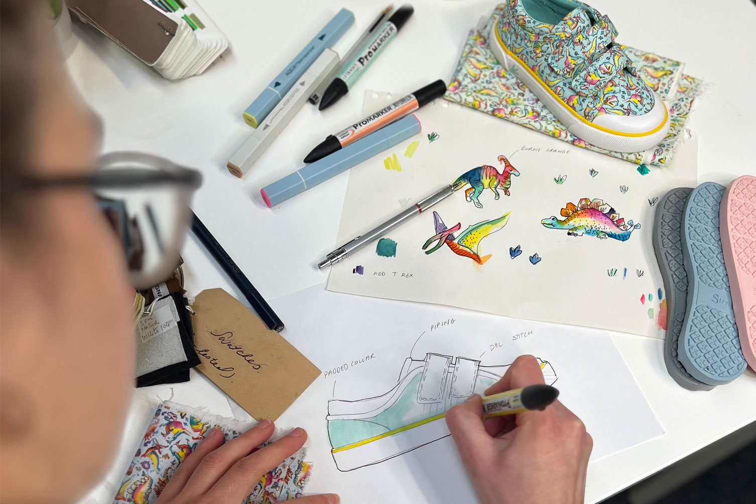 Designer colouring in a shoe sketch with pens, pencils and designs scattered around her