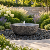 Blue Thumb 48-inch XL Natural Bowl Fountain with textured dark stone finish and bubbling water, surrounded by black polished pebbles in a professionally landscaped garden.