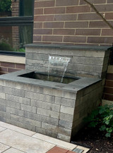 Small retaining wall waterfall cascading from a stainless steel spillway into a gray stone block water feature beside a brick building