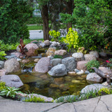 Small backyard garden pond with a rock waterfall, orange fish, boulders, and lush landscaping.