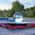 Crystal Sphere Fountain Kit - A reflective acrylic sphere fountain surrounded by red flowers, adjacent to a pool. Lush green trees in the background under a partly cloudy sky.