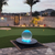 A serene backyard at dusk features a glowing blue acrylic crystal sphere fountain on a stone patio, adjacent to a putting green with lighted markers, framed by palm trees and a lit stone wall.