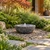 Blue Thumb 36-inch Natural Bowl Fountain with textured dark stone finish and gentle bubbling water, surrounded by mixed polished pebbles, Japanese maple, and lush garden landscaping.
