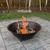 A large metal sugar kettle fire pit with bright orange flames burning logs is set on a stone patio. Behind, there's a grassy area and a forest with autumn foliage.
