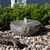 Natural stone swirl bowl fountain with water bubbling from the center, surrounded by mixed polished pebbles in a landscaped garden.