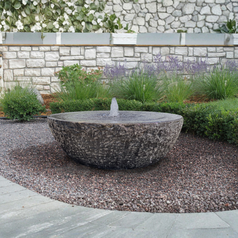A round stone fountain with softly bubbling water sits in a gravel bed. Behind, lush green plants and a stone wall create a serene garden setting.