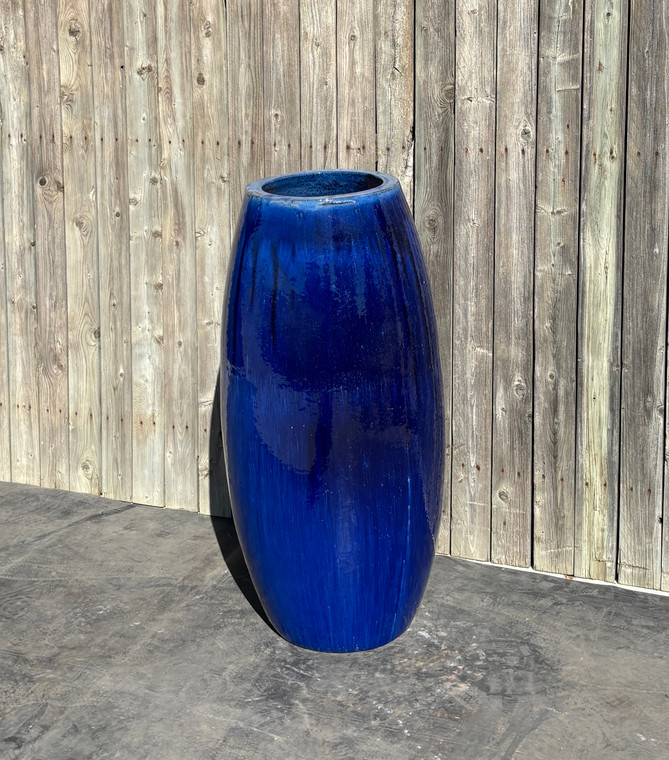 Tivoli Fountain Vase FNT30130 - A tall, glossy cobalt blue vase stands on a gray concrete surface against a rustic wooden fence. The vase's sleek design contrasts with the textured wood.