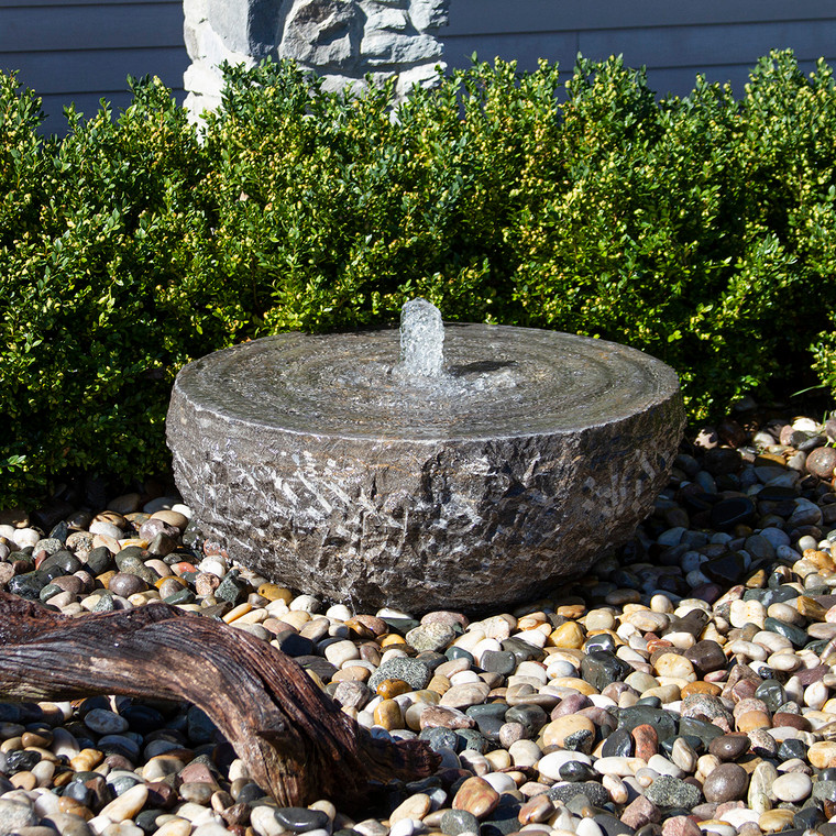 Natural stone swirl bowl fountain with water bubbling from the center, surrounded by mixed polished pebbles in a landscaped garden.