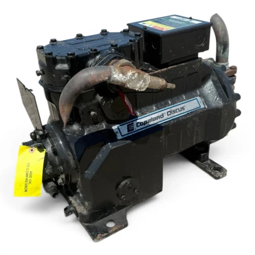 Front three-quarter view of the used Copeland 3DS3-R17MO-TFC 15 HP compressor (3 phase 208-230 volt), a black Copeland Discus unit with a yellow "ADD OIL TO COMPRESSOR" tag and a red round tag hanging from the valve, photographed with a second smaller black compressor visible in the background.