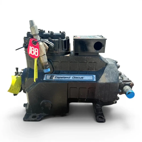 Front-left view of the used Copeland 3DS3-R17ME-TFC 15 HP compressor showing a copper fitting stub at upper right, the terminal box with a caution label at front left, the blue "Copeland Discus" nameplate across the housing, and a yellow oil-service tag hanging at left.