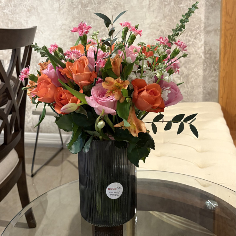 Bloomgol floral arrangement featuring peach and pink roses, carnations, and eucalyptus in a dark grey ribbed glass