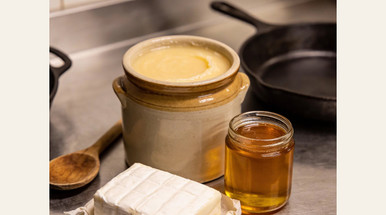 Three traditional cooking fats on a stainless steel counter - beef tallow in a crock, lard in butcher paper, and ghee in a jar