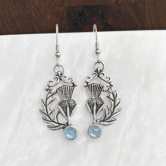 The Cirsium Gem Earrings feature thistle flowers, flanked by a curving leaf on one side, a 6mm gemstone at the bottom and beads and curl accents at the top. Shown with Blue Topaz.