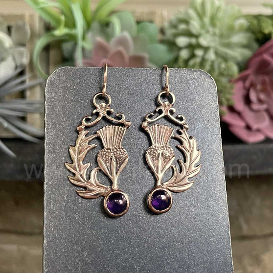 The Cirsium Gem Earrings feature thistle flowers, flanked by a curving leaf on one side, a 6mm gemstone at the bottom and beads and curl accents at the top. Shown in yellow gold with Amethysts.