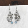 The Cirsium Gem Earrings feature thistle flowers, flanked by a curving leaf on one side, a 6mm gemstone at the bottom and beads and curl accents at the top. Shown with Blue Topaz.