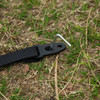 A black strap with a metal peg on grassy ground, designed for securing outdoor equipment.