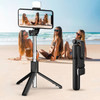 A black LED selfie stand with a smartphone mounted, set on a beach with three people in the background.