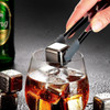 A drink glass filled with dark liquid and ice cubes, with a person's hand using tongs to place a metal ice cube.