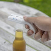 A drink bottle opener designed like a cap gun in white, held over a beer bottle on a wooden surface. It has a logo.