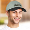 A grey and black cap is worn by a smiling person with blue eyes. The cap features a branding logo.