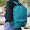 A teal backpack with a front pocket and mesh side pocket, worn on the back of a person. It has a logo.