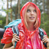 A woman wearing a transparent red emergency poncho carries a backpack in a forest setting, with trees in the background.