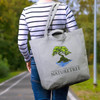 A grey tote bag featuring a tree design and a logo, carried by a person in a striped shirt on a pathway.