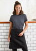 A woman wearing a black short waisted apron over a grey t-shirt, standing against a tiled wall. The apron features a logo.