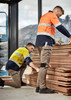 Two men in high-visibility work shirts and stretch pants, one crouching beside stacked timber, the other inspecting the wood.