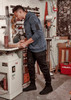 A man wearing a navy blue hoodie and black stretch jeans stands in a workshop, handling a piece of wood.