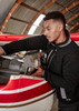 A man in a black fleece jacket works on an aircraft engine inside a hangar. The jacket features a logo.