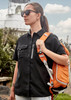 A woman in a black short sleeve outdoor shirt carries an orange backpack, standing near a lighthouse.