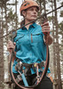 A woman in a blue outdoor short sleeve shirt, wearing a helmet and harness, stands on a zip line in a forest.