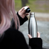 A drink bottle with a sleek stainless steel finish and a black lid, being held by a person outdoors.