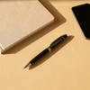 A silver ballpoint pen rests beside a notebook and a smartphone on a beige surface. The pen has a logo.