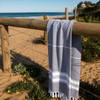 A grey and white Turkish towel draped over a wooden fence near a sandy beach and ocean.
