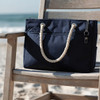 A navy waterproof beach bag with rope handles rests on a wooden chair by the shore. Features a logo.