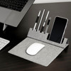A grey foldable mouse pad with a phone holder and slots for pens, placed beside a laptop and keyboard.
