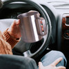 A stainless steel coffee mug with a handle, held in a hand inside a car. The mug has a logo on its side.