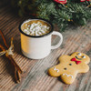 A white enamel camping mug filled with hot chocolate and marshmallows, alongside a gingerbread cookie and festive decorations.