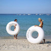 Two women on a beach holding large white inflatable swim rings, with clear blue water and boats in the background.