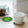 A green and brown custom PVC coaster sits on a wooden surface, with a white cup placed on top. The coaster features a logo.