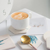 A white coffee cup on a hexagonal water absorbent coaster with blue floral designs, accompanied by a gold teaspoon and art print.