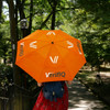 An orange sports umbrella with a logo, held by a person walking through a lush, green park setting.